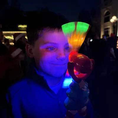 A young boy smiles as his face is illuminated by a spinning, light-up toy wand he is holding. The scene takes place outdoors at night, with blurred holiday lights and a crowd visible in the background.