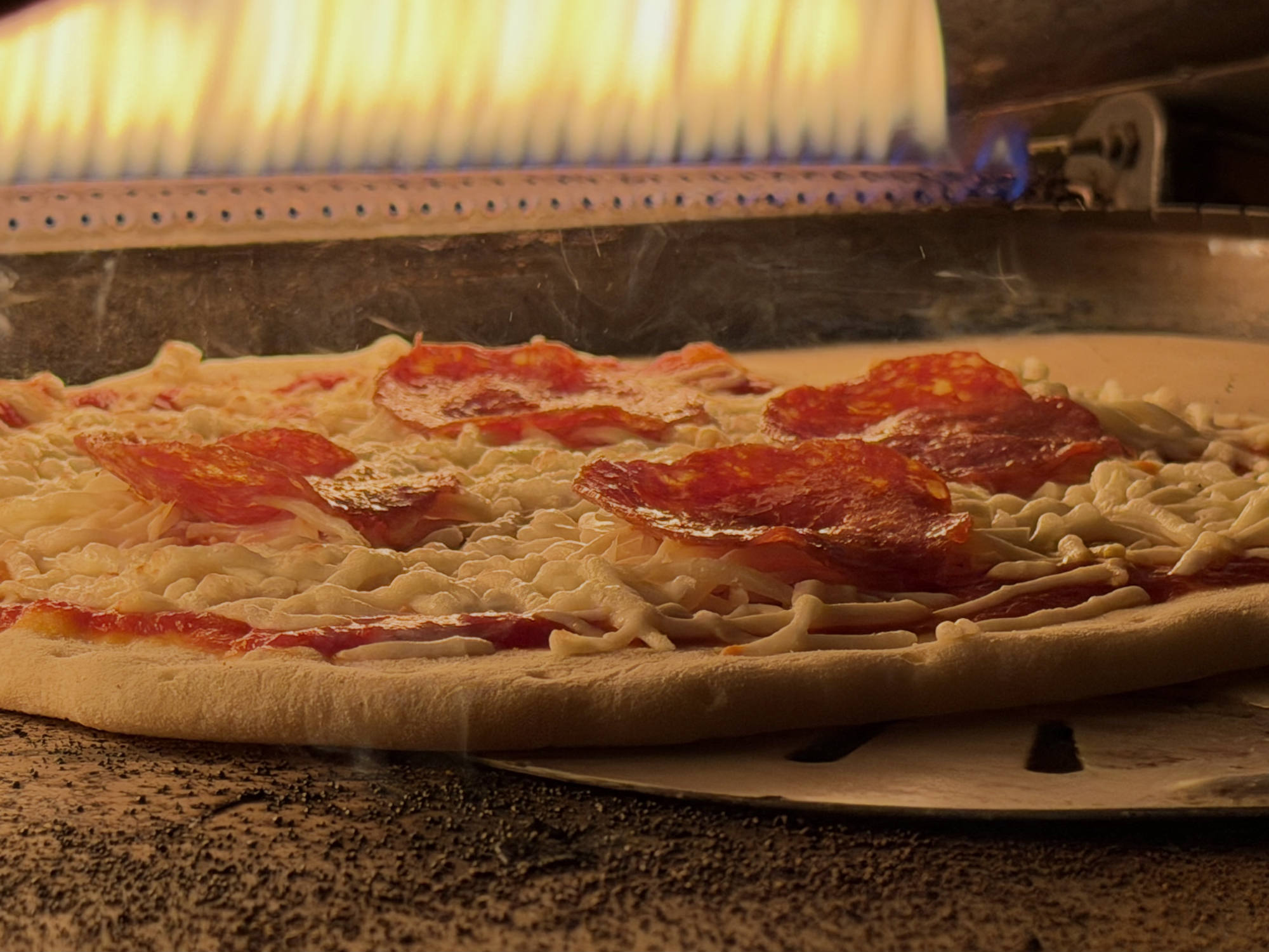 A close-up shot shows a pepperoni pizza baking inside a gas-fired oven, with flames visible in the background. The pizza sits on a metal peel over a textured stone surface as the cheese begins to melt.