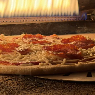 A close-up shot shows a pepperoni pizza baking inside a gas-fired oven, with flames visible in the background. The pizza sits on a metal peel over a textured stone surface as the cheese begins to melt.