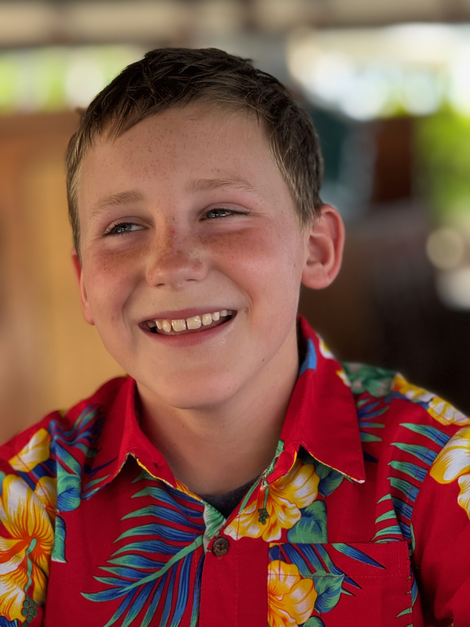 A close-up portrait of a smiling boy with freckles wearing a red floral Hawaiian shirt. He is looking off-camera against a softly blurred, warm-toned background.