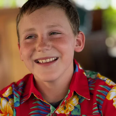 A close-up portrait of a smiling boy with freckles wearing a red floral Hawaiian shirt. He is looking off-camera against a softly blurred, warm-toned background.