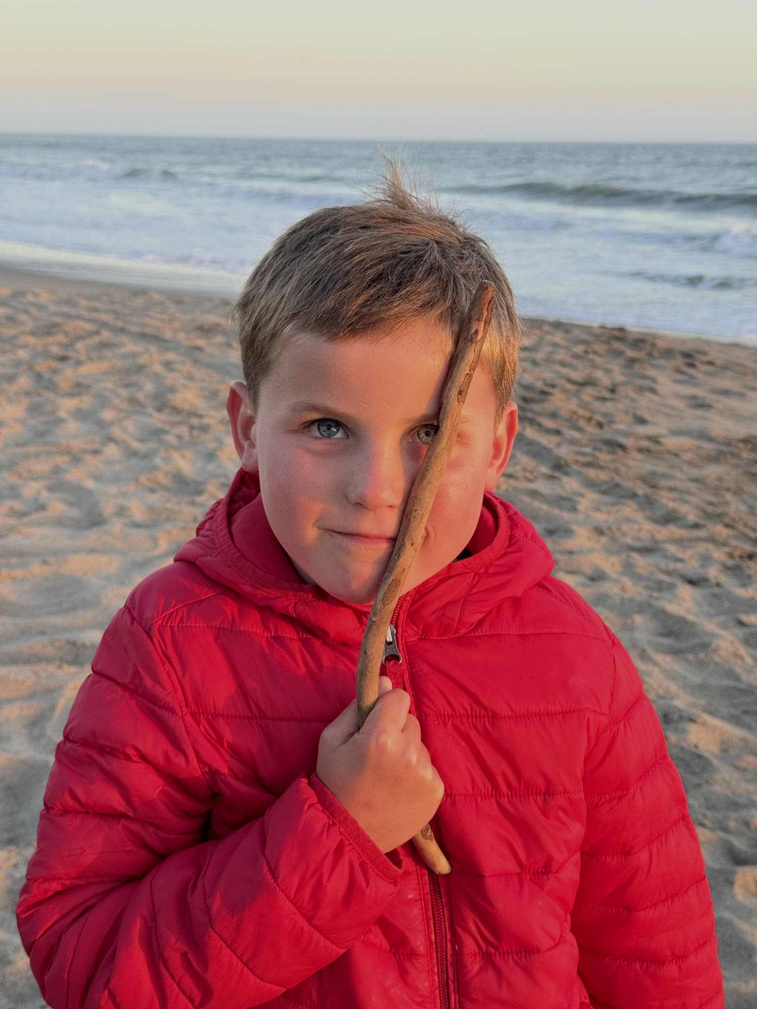 A young boy wearing a red puffer jacket stands on a sandy beach at sunset, holding a small stick vertically in front of his face. The ocean waves are visible in the background under a soft, warm sky.