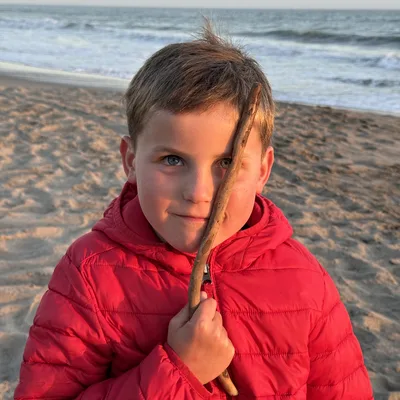 A young boy wearing a red puffer jacket stands on a sandy beach at sunset, holding a small stick vertically in front of his face. The ocean waves are visible in the background under a soft, warm sky.