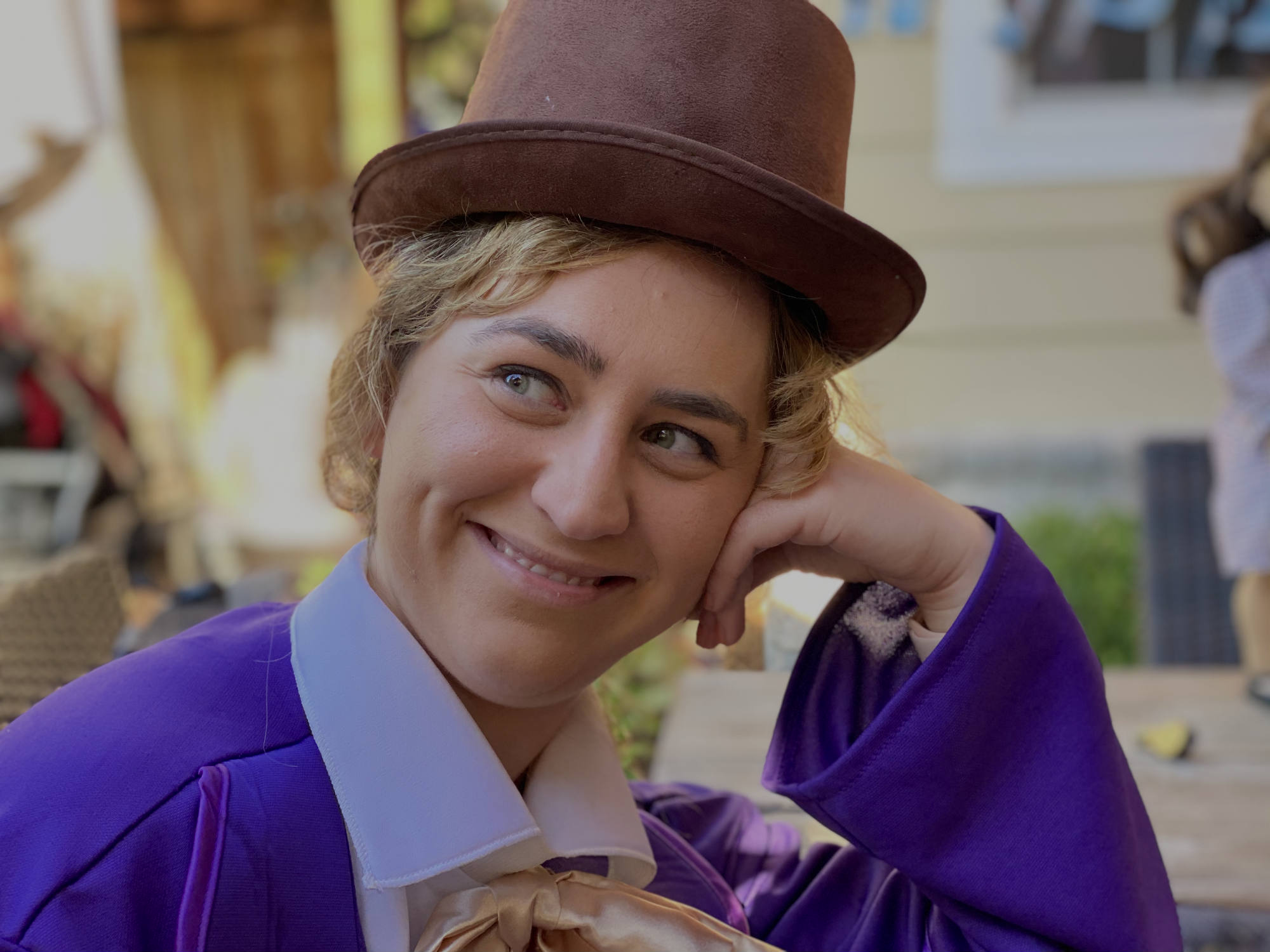 A close-up shot shows a smiling person wearing a Willy Wonka costume, complete with a brown top hat and purple coat, resting their head on their hand. They are outdoors in a blurred setting with buildings and seating visible in the background.