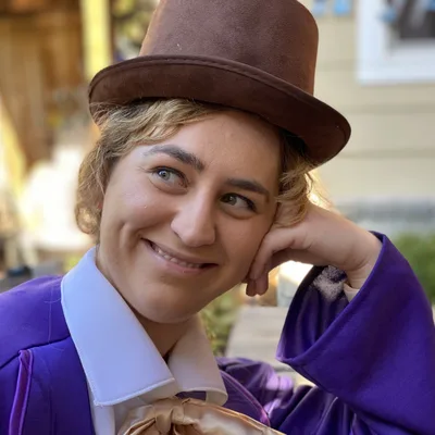 A close-up shot shows a smiling person wearing a Willy Wonka costume, complete with a brown top hat and purple coat, resting their head on their hand. They are outdoors in a blurred setting with buildings and seating visible in the background.