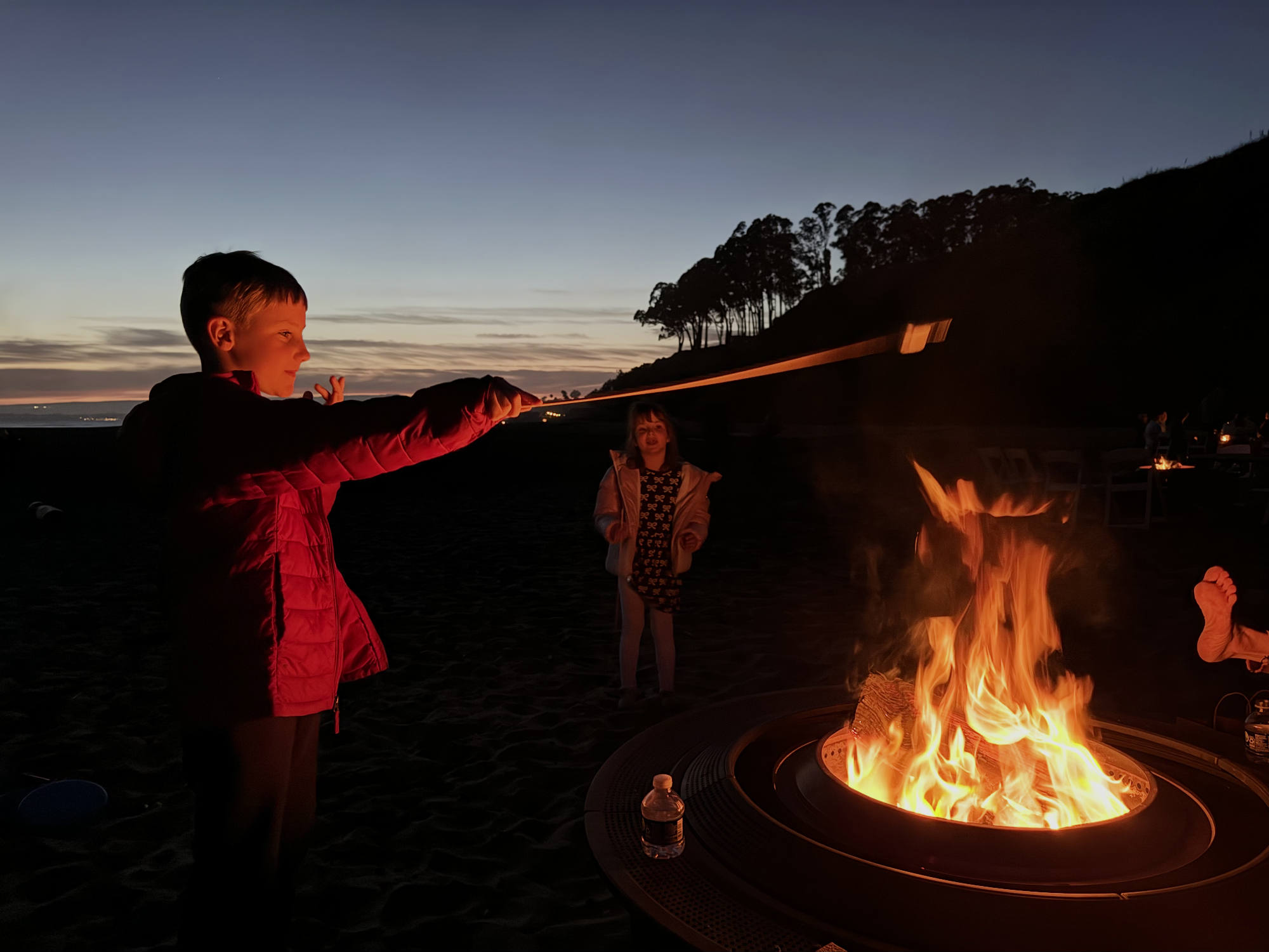 At dusk on a sandy beach, a boy in a red jacket holds a long stick over a blazing fire pit, while a girl stands in the background. The scene is illuminated by the warm glow of the fire, with a silhouetted treeline and a sunset sky visible in the distance.