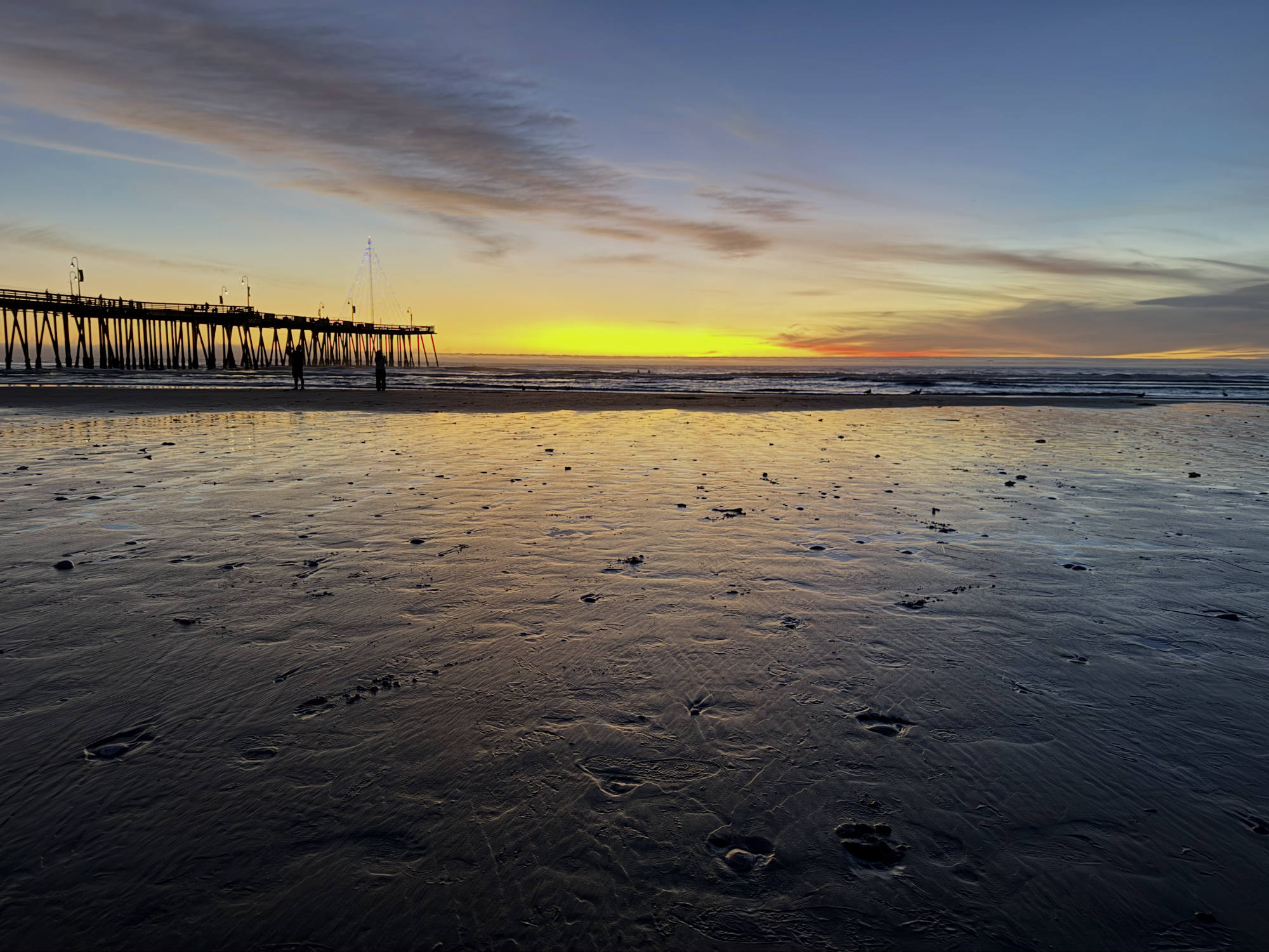 A wide-angle shot captures a sunset over the ocean, with a long wooden pier extending into the water on the left and a wet, sandy beach in the foreground reflecting the yellow and blue sky. Two silhouetted figures stand near the shoreline, and a lit, tree-shaped light display is visible on the pier.