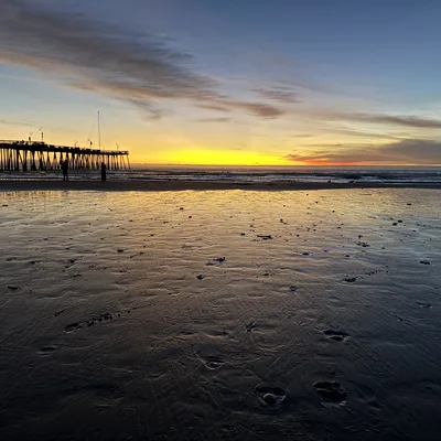 A wide-angle shot captures a sunset over the ocean, with a long wooden pier extending into the water on the left and a wet, sandy beach in the foreground reflecting the yellow and blue sky. Two silhouetted figures stand near the shoreline, and a lit, tree-shaped light display is visible on the pier.