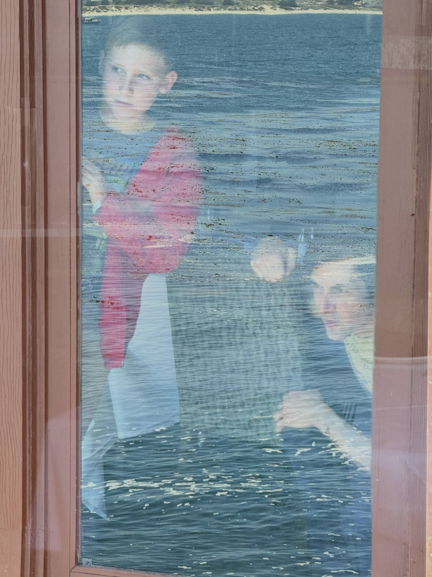 A boy and a woman are seen through a window pane that reflects a body of water, creating a double-exposure effect. They appear to be looking out from inside a building toward the water.