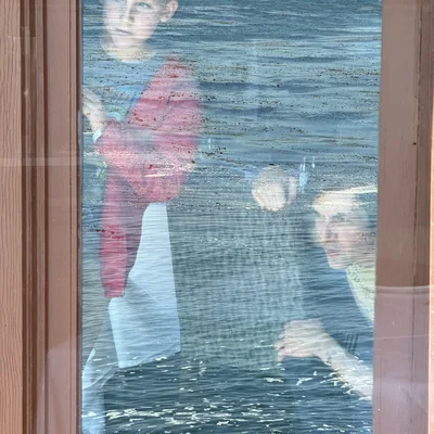 A boy and a woman are seen through a window pane that reflects a body of water, creating a double-exposure effect. They appear to be looking out from inside a building toward the water.
