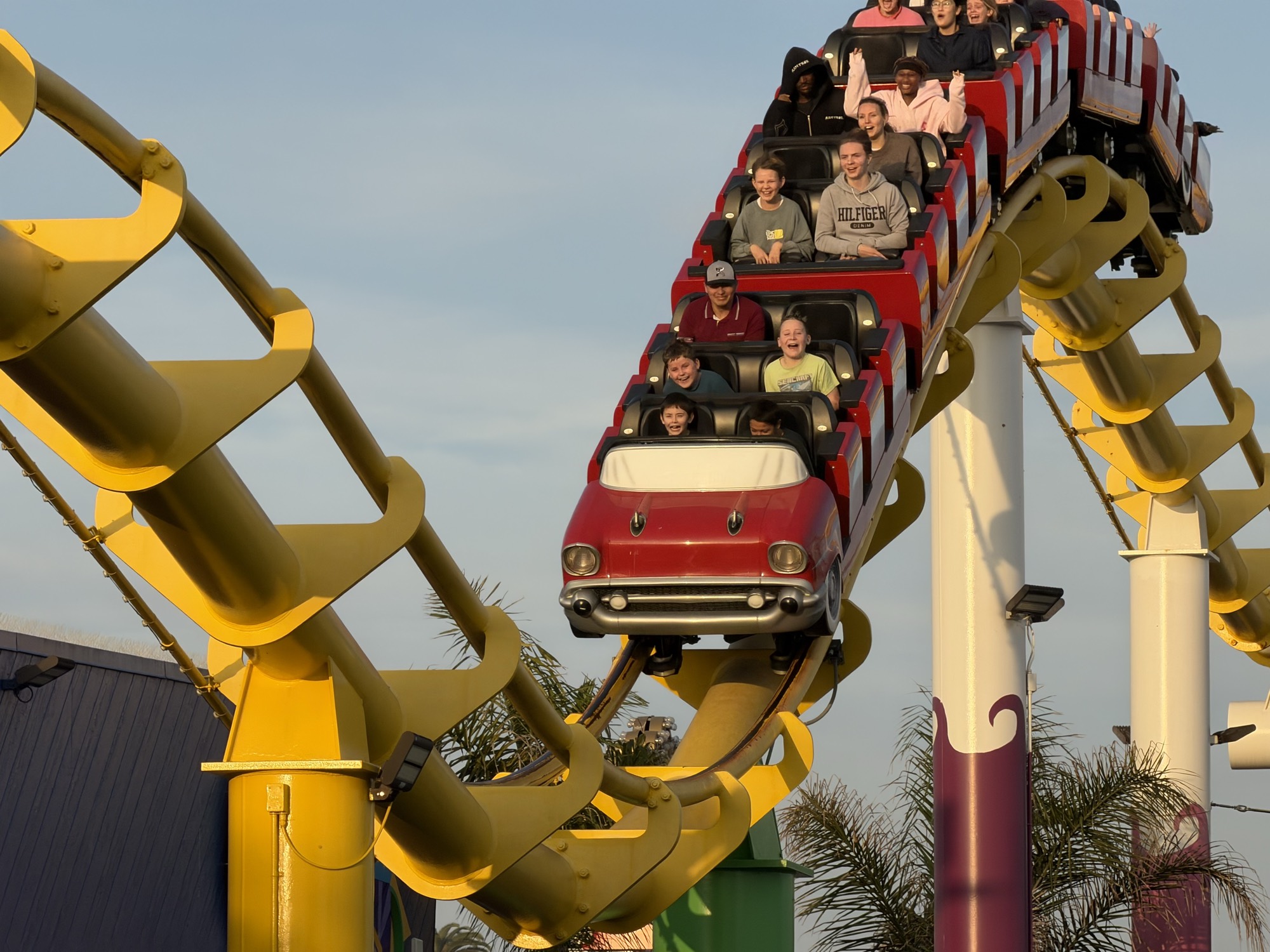 A group of people ride a roller coaster with a red, car-shaped front car on a bright yellow track. The outdoor amusement park ride is captured against a clear blue sky during the day.