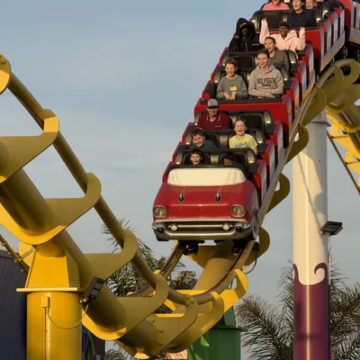 A group of people ride a roller coaster with a red, car-shaped front car on a bright yellow track. The outdoor amusement park ride is captured against a clear blue sky during the day.