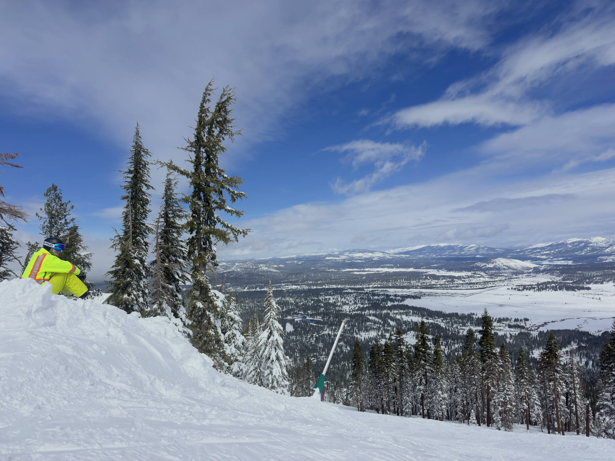 A person in a bright yellow ski jacket sits on a snowy ridge, looking out over a vast, snow-covered valley with pine forests and distant mountains under a blue sky with wispy clouds.