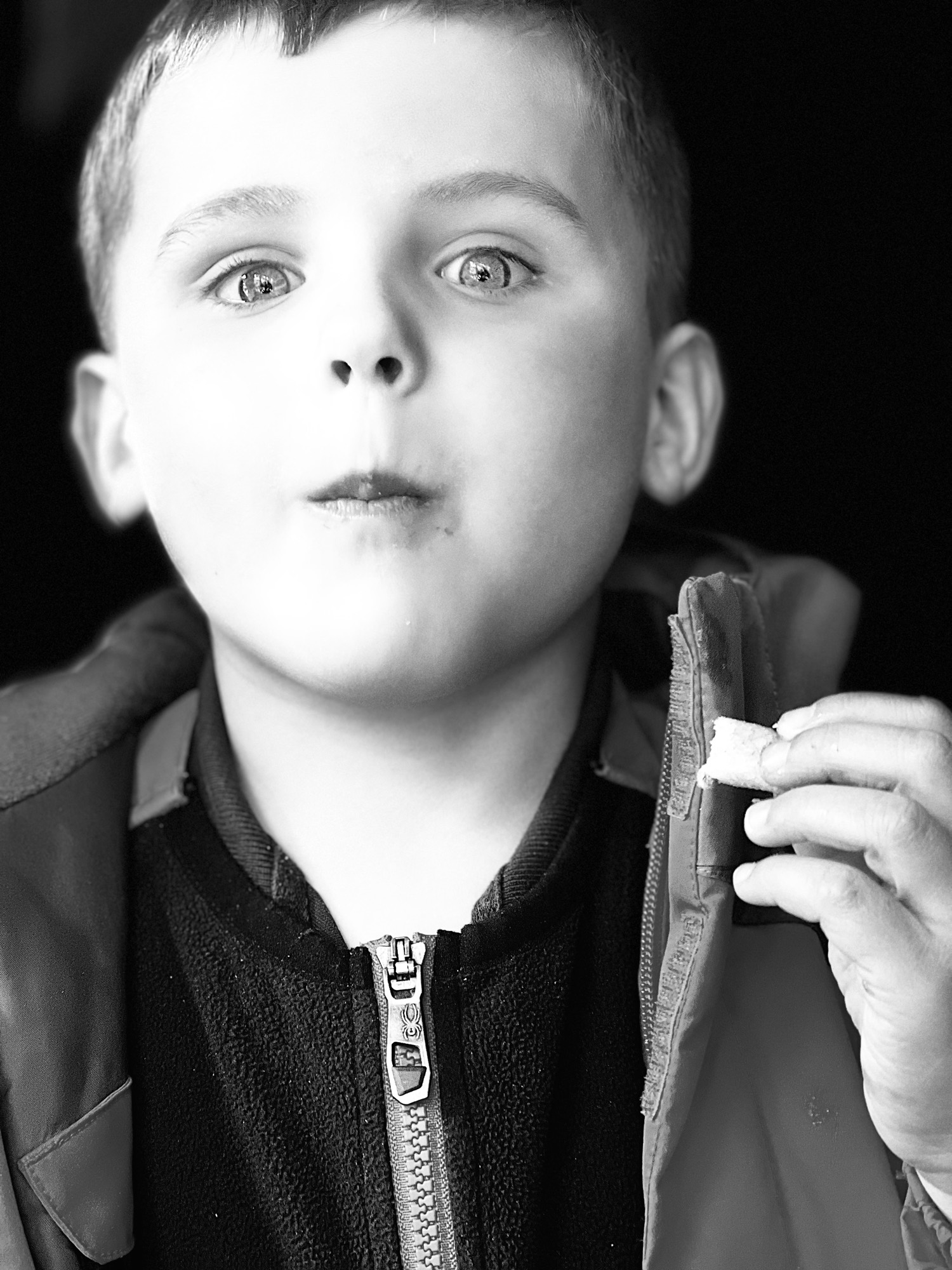 In this black and white close-up portrait, a young boy wearing a zippered jacket looks directly at the camera with wide eyes while holding a small piece of food in his hand. The background is a solid dark color, focusing all attention on the child's expression.