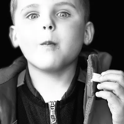 In this black and white close-up portrait, a young boy wearing a zippered jacket looks directly at the camera with wide eyes while holding a small piece of food in his hand. The background is a solid dark color, focusing all attention on the child's expression.